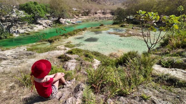 Woman in a hat and skirt relaxing on a large rock, observing the vibrant turquoise-green water of Wadi Darbat, a natural park oasis in Taqah, Dhofar Governorate, Oman