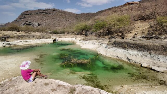 Woman in a hat and skirt relaxing on a large rock, observing the vibrant turquoise-green water of Wadi Darbat, a natural park oasis in Taqah, Dhofar Governorate, Oman