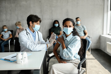 Middle-eastern woman patient in protective face mask getting vaccinated against COVID-19 at clinic. Multiethnic group of people waiting for immunization, male doctor making injections