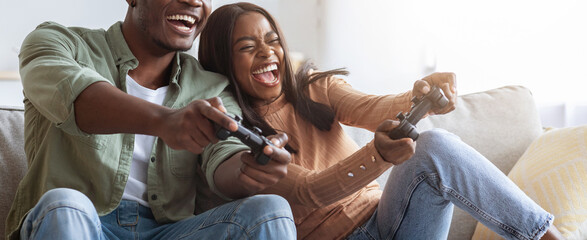 A couple is joyfully playing video games together on a comfortable couch in their living room. They are deeply focused and laughing while holding game controllers, enjoying each other's company.