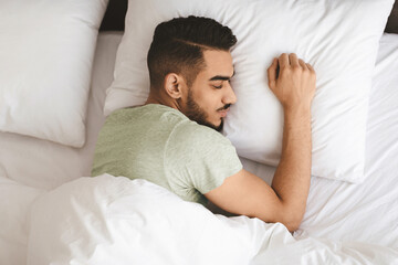 A young man with a trimmed beard is resting comfortably in bed, lying on his side with his arm gently hugging a pillow. The setting is calm, featuring white bedding and a serene atmosphere.