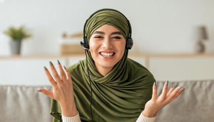 A young woman with a green headscarf and headphones smiles brightly while gesturing with her hands, showing excitement during an online conversation in a cozy room.