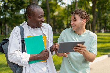 Two young men are smiling and engaging in a lively discussion in a park. They are surrounded by greenery while using a tablet and holding notebooks, enjoying their time together.