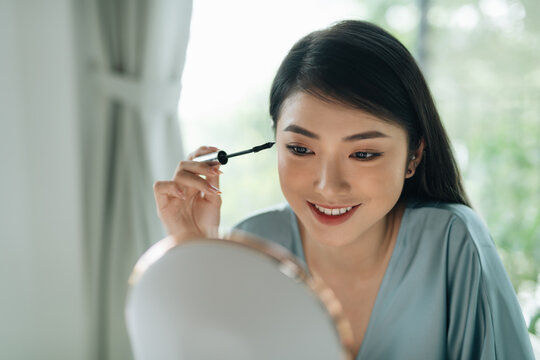 Happy young lady applying mascara on her lashes near mirror 