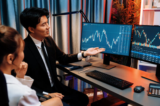 Asian businessman showing dynamic stock exchange currency rate statistic online to share with coworker, explaining to compare with high or low on computer screen at night neon modern office. Infobahn.