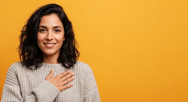 Smiling young adult woman with dark wavy hair placing hand on chest, expressing gratitude and sincerity, isolated on a vibrant orange background.