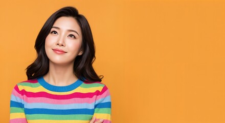Happy young Asian woman in a vibrant rainbow striped sweater looking up with a dreamy expression, isolated on orange background.
