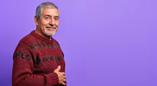 Happy senior Hispanic man with a grey beard and patterned red sweater smiling confidently against a vibrant purple studio background