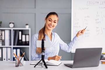 A teacher is delivering an online English lesson in a cozy classroom. She is smiling and pointing at a whiteboard filled with notes.