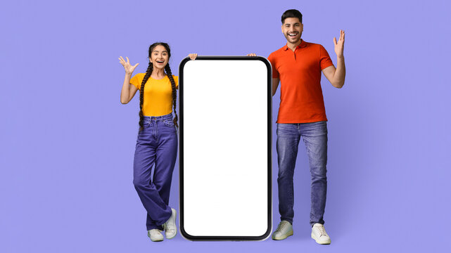 Two young adults are joyfully posing together, holding a large blank smartphone. They are dressed casually with a vibrant purple background, creating a lively and engaging scene.