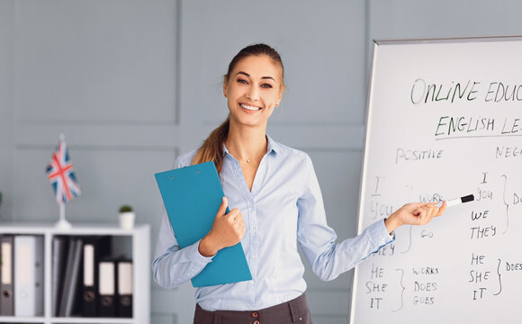 A smiling teacher stands next to a whiteboard, explaining English language concepts during an online class. Her welcoming attitude and clear instruction encourage active student participation.