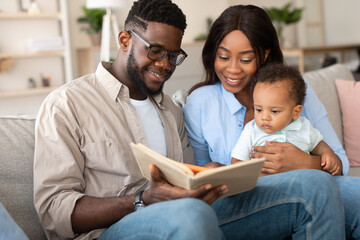 Millennial black parents joyfully read a book to their toddler boy while relaxing on the couch in...