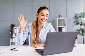 A woman with long hair is sitting at a desk, smiling and waving at her laptop during a virtual...