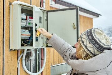 Mature woman checks electricity meters mounted on yellow wooden siding house wall.