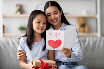 A young Asian woman joyfully reads a handmade Mother's Day card while sitting on a couch with her...