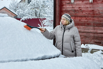 Brushing removes deep snow from car roof as woman in warm winter jacket and knit hat clears vehicle...