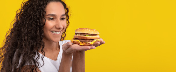 A woman with curly hair smiles broadly while holding a delicious, large burger in her hands. The...