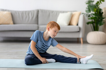 Cute redhead boy sits on fitness mat at home, stretching while reaching for his foot. The living room is cozy with soft furniture and plants around, creating a calm atmosphere for exercise.