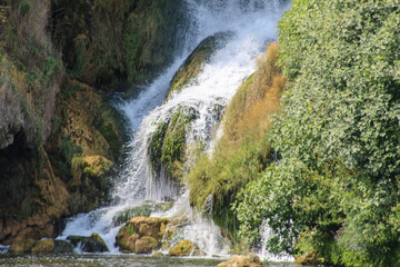 A close-up view of the Kravica Waterfall in Herzegovina, showing cascading water over moss-covered rocks surrounded by dense green vegetation.