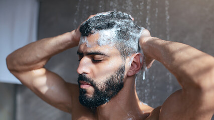 A man is showering and applying shampoo to his hair, focusing on self-care in his bathroom. Water cascades down, creating a relaxing atmosphere for his grooming routine.