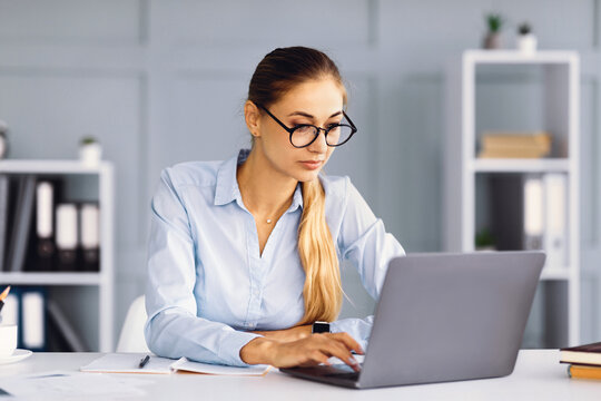 A woman with long hair and glasses is typing on her laptop at a tidy desk in a bright home office. The shelves behind her display books and decorative items, creating a professional atmosphere.