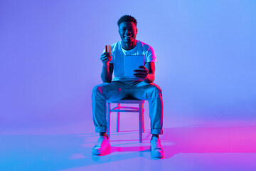 A man sits on a chair, smiling while holding a tablet in one hand and a card in the other. The...