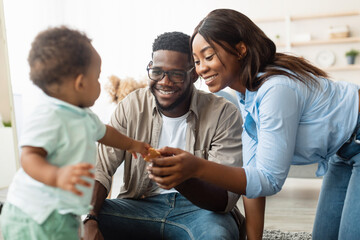 Cheerful millennial parents play with their toddler son at home. The family sits on a carpet...