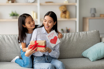 A daughter joyfully surprises her mom with a handmade card and gift box on Mother's Day. They sit...