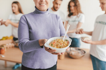 A woman smiles while holding a bowl of colorful food in a kitchen where friends are engaged in cooking. The atmosphere is friendly and lively as they share food.
