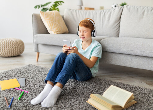A cheerful preteen redhead boy sits on a carpet at home, wearing a wireless headset and focused on his smartphone. He is doing homework while surrounded by school supplies and a book.