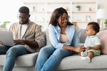 In a bright living room, a black mother sits on the couch, happily playing with her baby. Meanwhile, the father focuses on his laptop, balancing work and family time in their home.