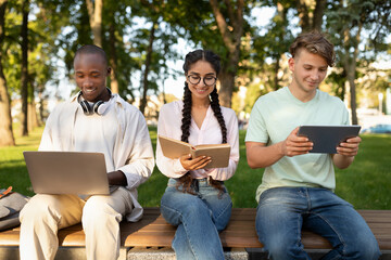 Three young adults sit on a bench in a park, each focused on their devices. One uses a laptop,...