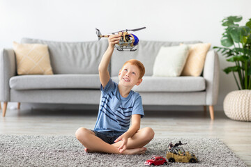 In a bright living room, a cheerful redhead boy sits on a carpet playing with a toy helicopter and...