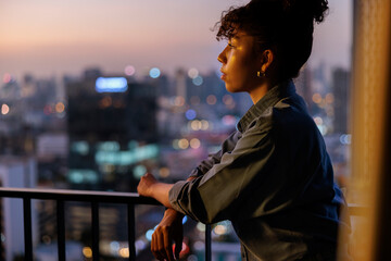 Woman gazes at city lights from balcony at dusk.