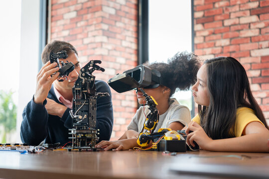 Mentor Guiding Students in Robotics and Virtual Reality (VR) Class, Man Showing Robotic Component to Girls with VR Headset, Children Learning Engineering and Programming with Adult Supervision - Powered by Adobe