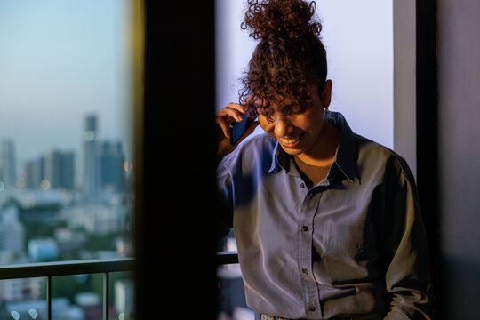 Woman smiles on balcony, city lights blur in background.
