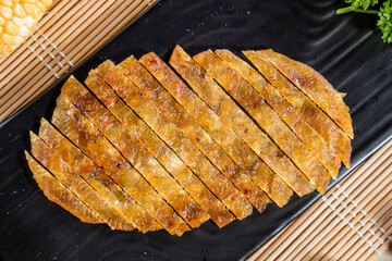 Sliced grilled tofu skin arranged on a black plate in a fan shape, with corn and parsley on bamboo mat background.