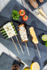 Skewers of okra, chicken, and beef topped with melted cheese, served with lime, lemon, tomato, parsley, and pinecones on slate board.