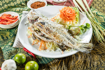 A whole grilled fish served on shredded cabbage and carrots, garnished with carved carrot flower and herbs, surrounded by Thai spices.
