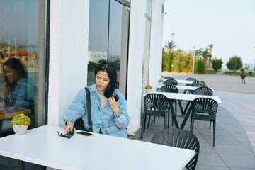 Woman at outdoor cafe sits beside a white table, holding sunglasses and glancing toward the storefront, denim jacket, casual mood, empty chairs, calm daytime urban dining setting