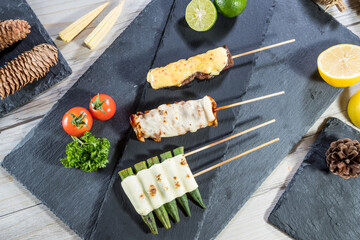 Skewers of grilled meat, chicken, and okra topped with melted cheese, served with lime, lemon, tomato, and pinecones on slate board.