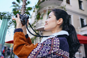 Woman photographer captures moment with camera on urban street, vibrant jacket and patterned sweater, lens focused on city life, daylight mood, candid style, creative storytelling in motion