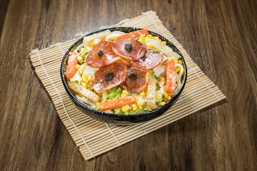 A colorful salad bowl with pepperoni, noodles, corn, lettuce, carrot, cheese, and black garnish.