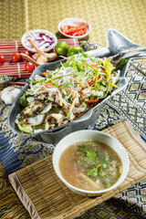 Thai-style fish served in metal dish with vegetables and herbs, accompanied by soup and Thai ingredients.