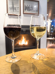 Two glasses of wine one white one red on a table in front of a warm cosy winter fireplace with orange flames