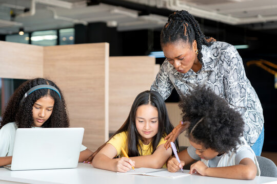 African American Mentor Guiding Diverse Students with Homework and Learning, Multicultural Teacher Helping Girls with Writing and Laptop Use in Modern Classroom