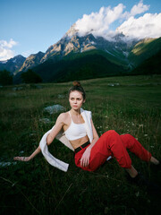 Fototapeta premium Woman resting on a meadow with mountains in the distance, wearing a white top and red pants, scarf over her shoulder, showcasing casual outdoor fashion and natural scenery