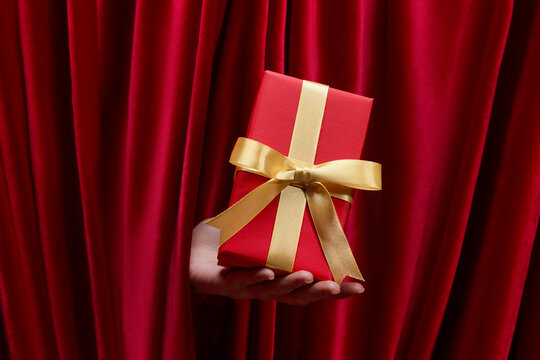 Hand holding a red gift box with a gold ribbon against a red curtain.