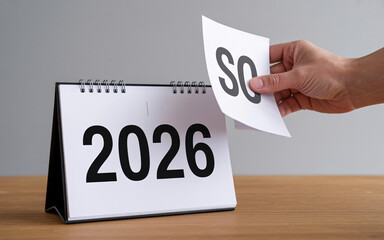 A hand turning a page on a calendar showing the year of two thousand and twenty six on a wooden table