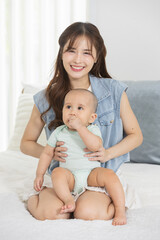 Smiling young Asian mother holding child boy on her lap in cozy home. Kid sucking hand shows bonding and love. Perfect for motherhood, parenting, baby care and family lifestyle themes.